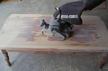 Using a belt sander to remove the finish and stain from a coffee table top.